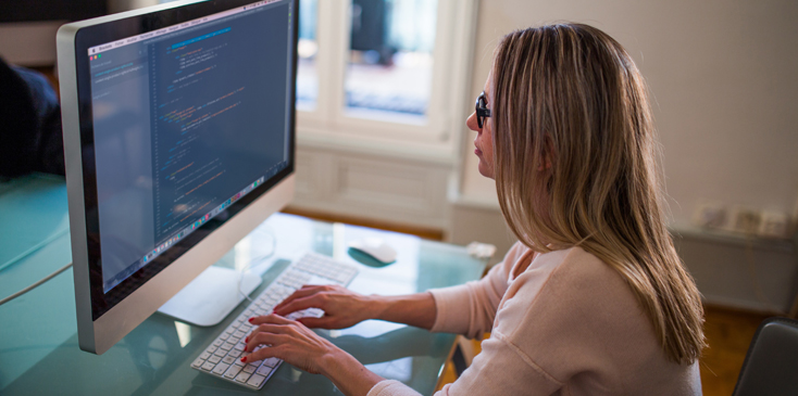 A woman with blonde hair wearing a pink sweater works on an iMac computer