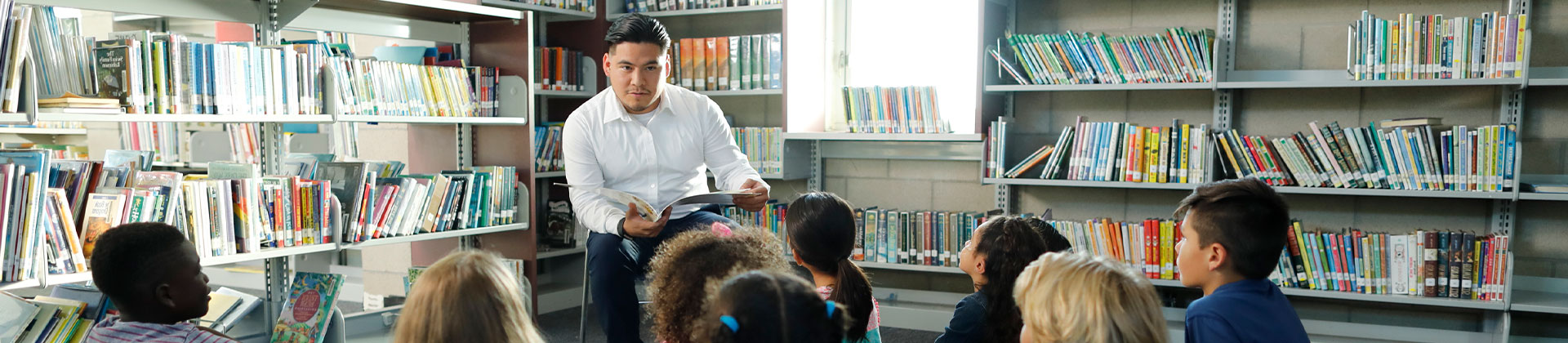 A man in a white dress shirt sits in a corner of a library and reads a story to young children, who are seated on the ground in front of him.