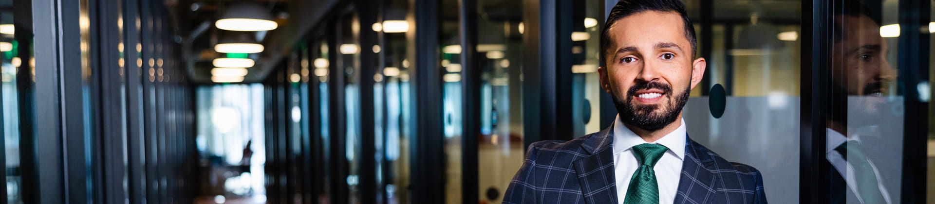 Ivan F. stands in a long glass-walled hallway. He wears a plaid navy suit 