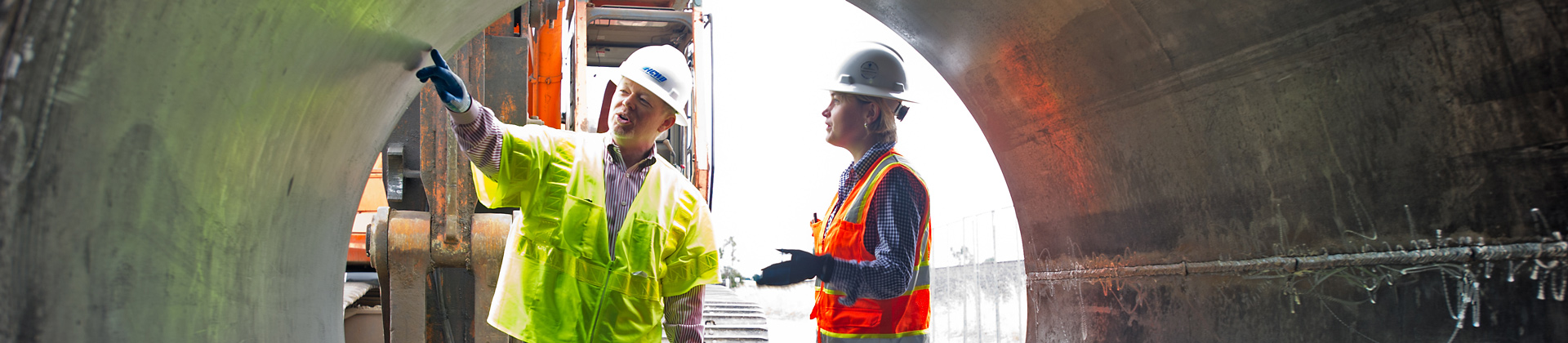 A man and a woman in hard hats and safety vests look at a giant pipe