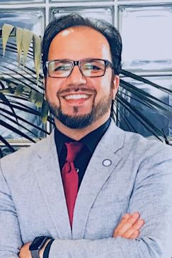 Monir Masoud crosses his arms while wearing a light gray suit and red tie