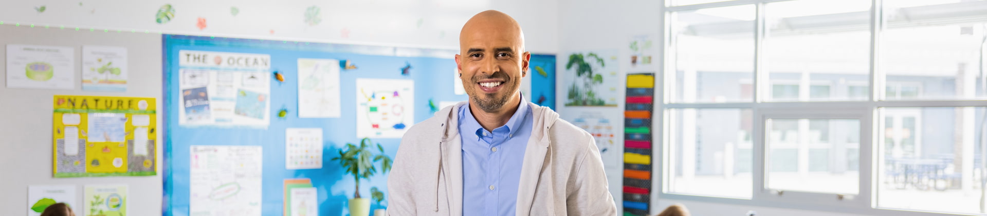 A bald man with earrings and a white hoodie stands in a classroom