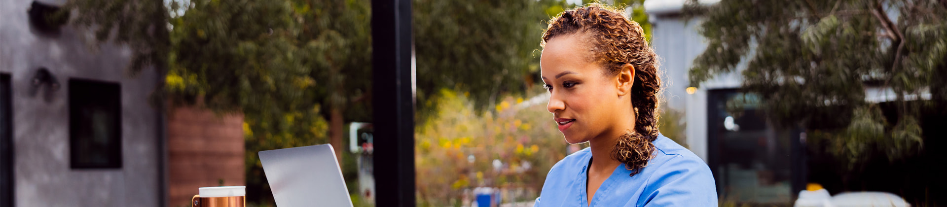 A woman in blue scrubs sits outside looking at a laptop