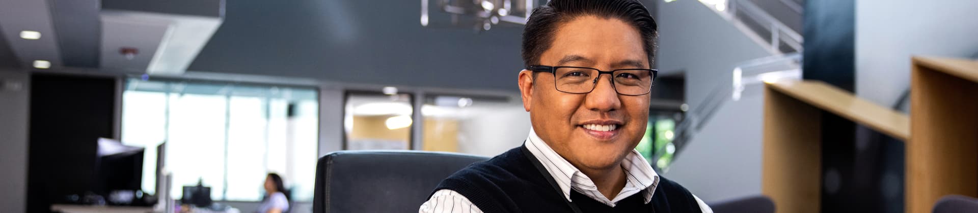 Man wearing glasses and a sweater vest sits in a chair smiling.