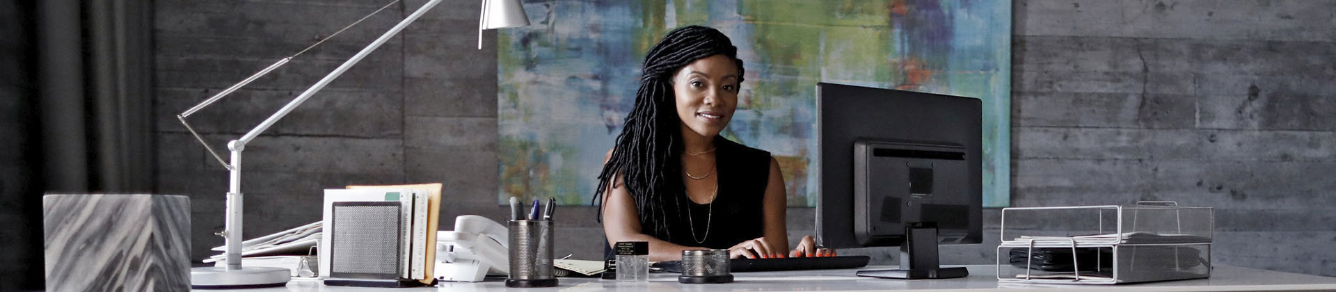 Woman with long microlocs sits at a desk smiling with hands on keyboard