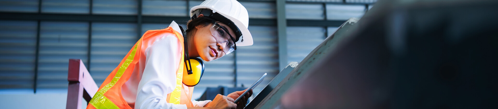 woman in hard hat, safety glasses and orange vest looks at machinery