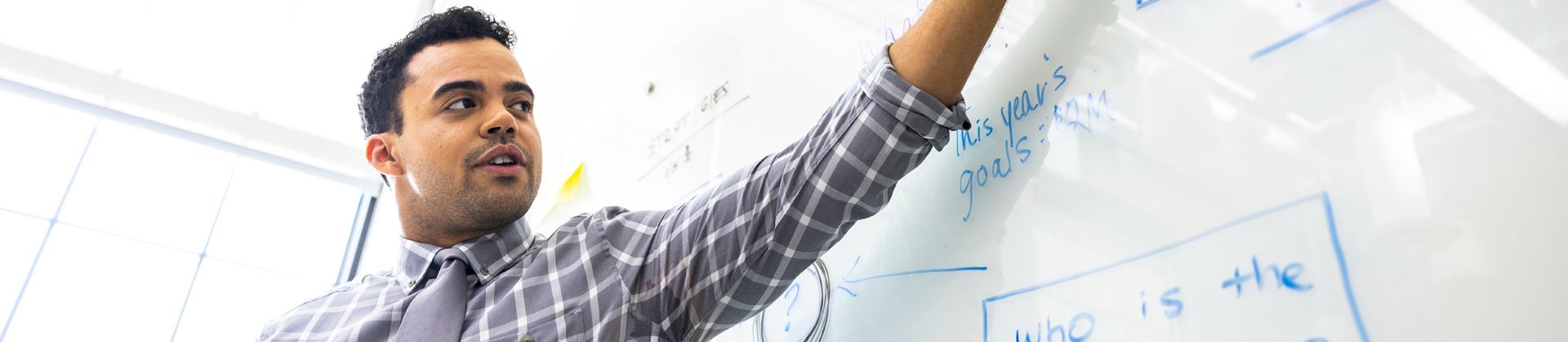 Man in a dress shirt and tie teaches at a whiteboard. Minor in Business Studies Program Page