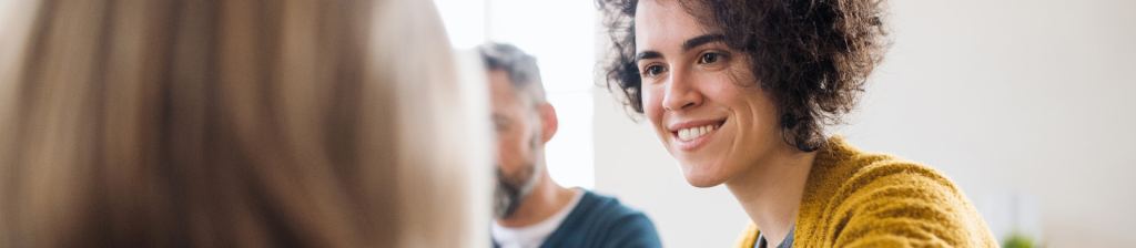 woman with short brunette hair looking to her left at woman with blonde hair, smiling at woman with man blurred in background