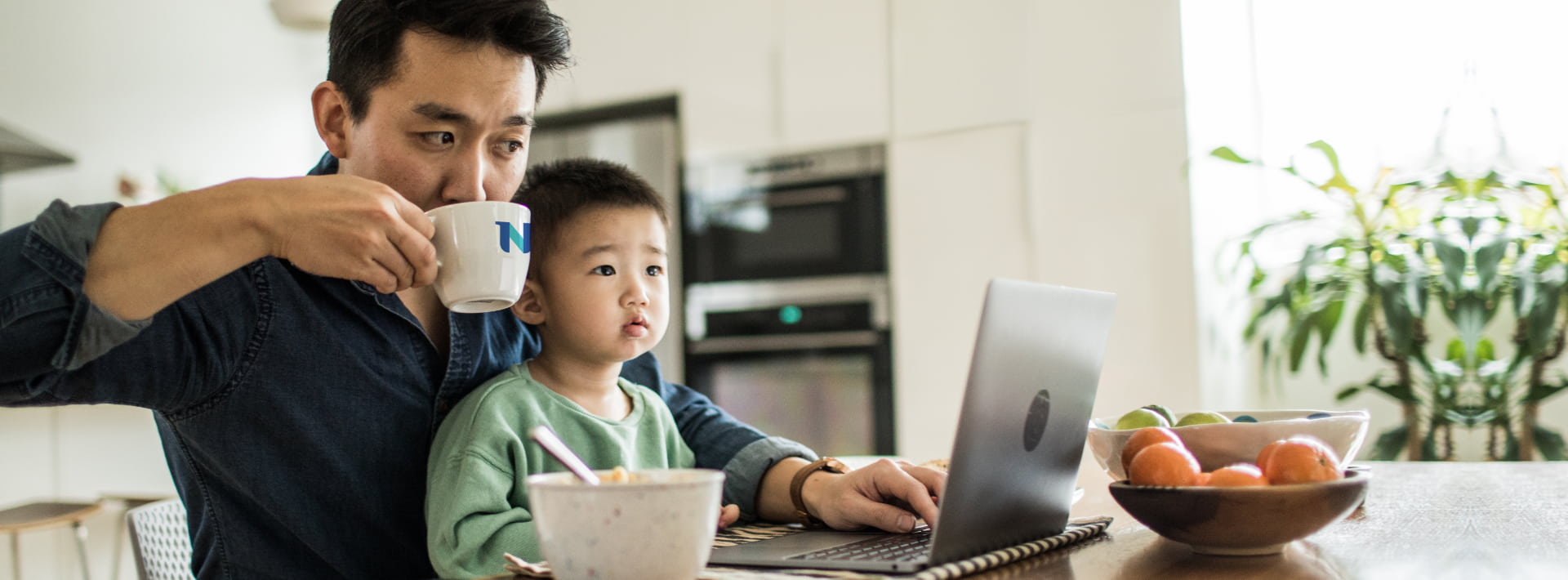 Father sipping mug as son sits on lap and looks at open laptop