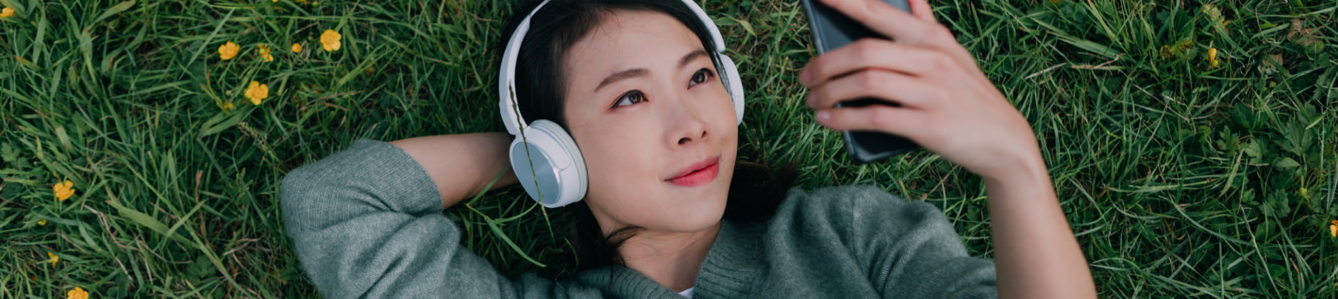 woman wearing headphones lies in the grass and holds her phone up