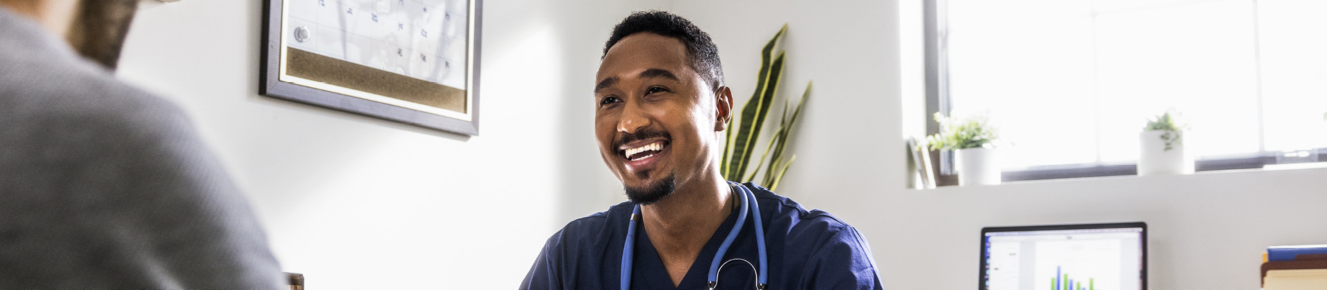 A man in scrubs sits in an office and meets with a patient, who is out of frame