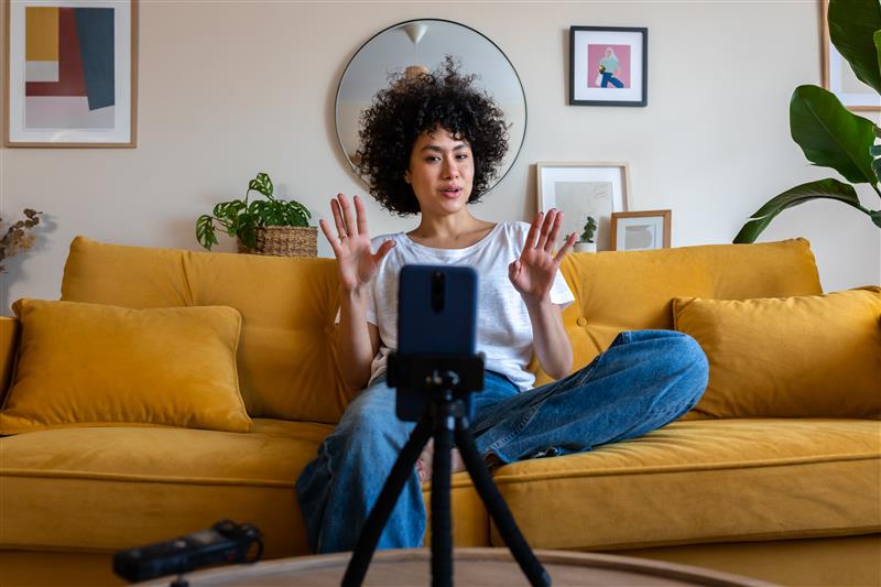 woman sitting on mustard colored sofa making a video with her phone